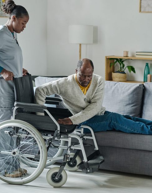 African young social worker helping man with disability to stand up, she caring about him at home